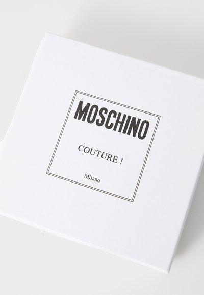 Balts taisnstūra kaste ar vāku, ar melnu Moschino logo un vārdu "COUTURE!" rakstītu lielajiem burtiem, apvilktu ar plānu melnu malu.