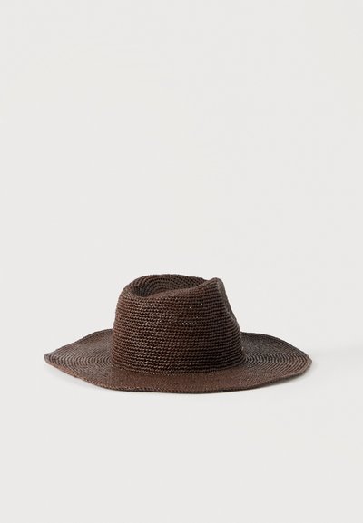 Chapeau en paille tressée marron à large bord avec une calotte plissée, posé sur une surface blanche plate.