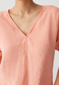 Pfirsichfarbene Bluse mit V-Ausschnitt, kurzen Ärmeln und einem weichen, strukturierten Stoff. Eine Mittelnähten zieht sich vertikal über die Vorderseite.