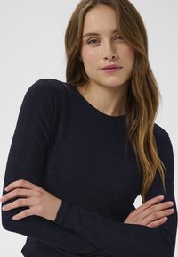 Námornícky modrý top s dlhým rukávom a priliehavým strihom, vyrobený z jemnej textúrovanej tkaniny, ktorá má jemný lesk po celom povrchu.