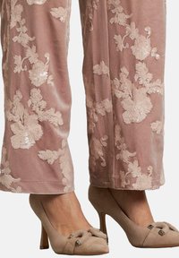 Primo piano di gambe che indossano pantaloni in velluto rosa con ricami fiorati in paillettes e scarpe con tacchi alti in suede nudo con fibbie decorative.