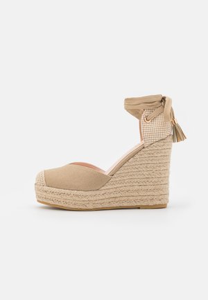 Beige kile-espadrillesandal med flettet platform, ankelbind og frynse detaljer.