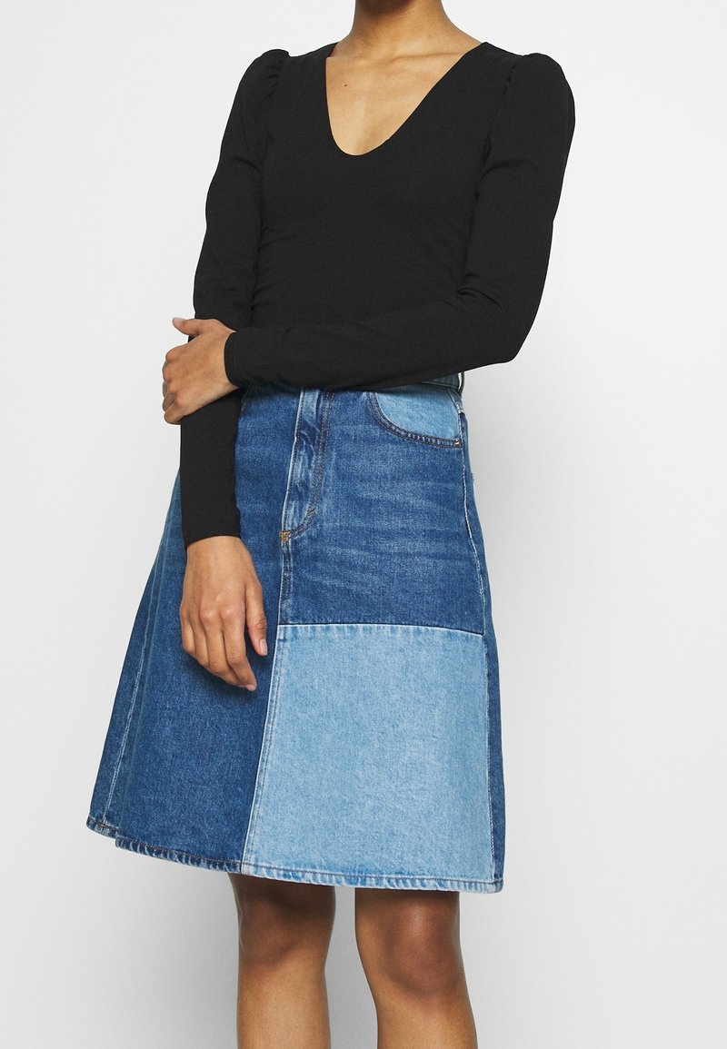 Schwarzes Langarm-Oberteil mit tiefem V-Ausschnitt, kombiniert mit einem knielangen Jeansrock, der aus einem Patchwork aus hellblauen und dunkelblauen Abschnitten besteht.