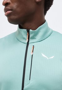 Κοντινό ενός ατόμου που φοράει ένα ανοιχτό τιρκουάζ μπουφάν με ψηλό γιακά και δύο μαύρα φερμουάρ με πορτοκαλί σημεία έλξης.