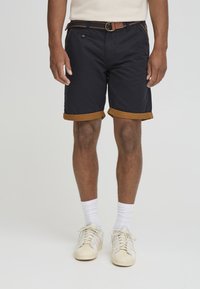 Mørkeblå shorts med brune oppbrett, frontlomme og beltehemper, parret med hvite sokker og kremfargede sneakers.