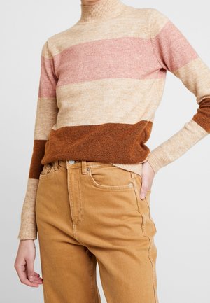 Suéter de punto con cuello alto, decorado con rayas horizontales en rosa, beige y marrón. Combinado con jeans de mezclilla color arena, que muestran los puños y los bolsillos.