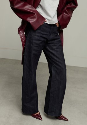 Donkerblauwe wijde jeans met een gladde textuur, gecombineerd met een grijze top en een diep bordeaux leren jas met metalen drukknoopjes.