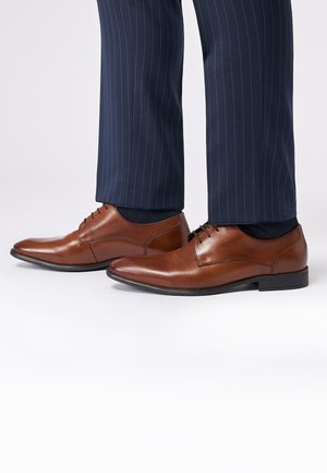 Brown leather dress shoes with a pointed toe, lacing detail, and a smooth texture. Paired with dark pinstriped trousers.