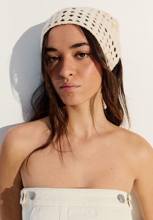 Mladá žena s dlouhými tmavými vlasy, která nosí béžovou háčkovanou šátek na hlavě a bílý top bez ramínek, stojící na světlém pozadí.