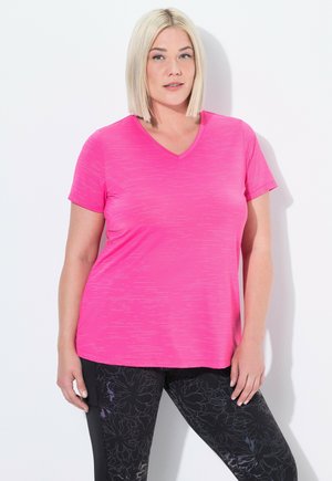 Žena s krátkými blond vlasy, která má na sobě světlou růžovou sportovní kratší tričko a černé legíny s květinovým vzorem, stojí proti bílé zdi.