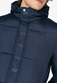 Marineblauwe gevoerde jas met hoge kraag, ritssluiting en geborduurde letters. Heeft een metalen knoop en logoaccent op de borst.