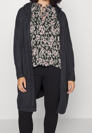 Femme portant un chemisier noir à fleurs, un long cardigan gris foncé côtelé avec capuche, et un pantalon noir ajusté, debout devant un fond uni.