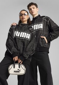 Jachete negre matlasate cu un model în grilaj alb și cu un logo „PUMA” proeminent; asortate cu o poșetă mică albă cu accente negre.
