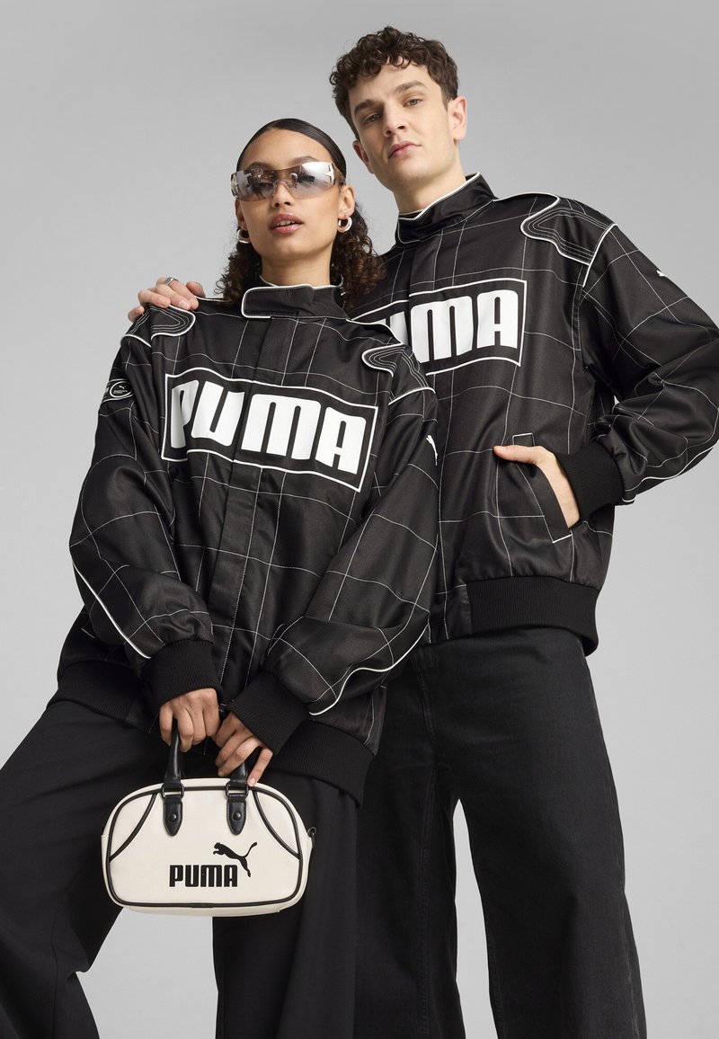 Jachete negre matlasate cu un model în grilaj alb și cu un logo „PUMA” proeminent; asortate cu o poșetă mică albă cu accente negre.