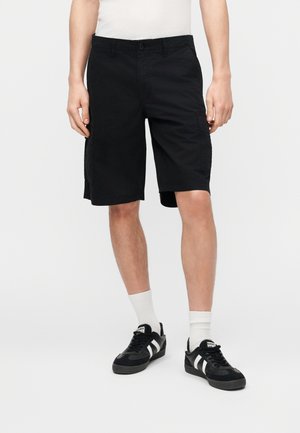 Hombre con pantalones cortos cargo negros, calcetines blancos y zapatillas negras con rayas blancas, de pie contra un fondo blanco liso.