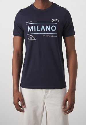 Mann trägt ein dunkelblaues Armani Exchange T-Shirt mit „MILANO“ und weiteren stadtbezogenen Texten, kombiniert mit off-weißen Hosen.