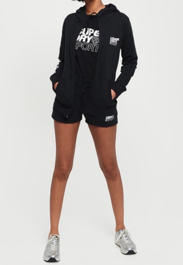 Sudadera negra con cremallera sobre una camiseta negra con estampado blanco, combinada con pantalones cortos negros con cordones y zapatillas deportivas plateadas.