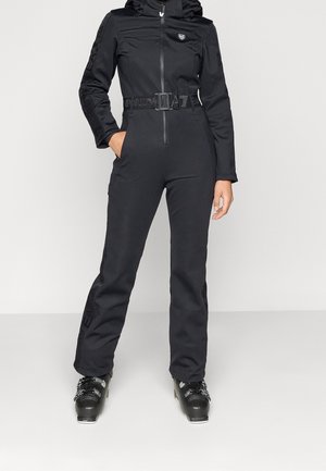 Femme portant une combinaison de ski noire ajustée avec une fermeture éclair à l'avant, une ceinture et des chaussures de ski, debout avec une main dans la poche sur un fond uni.