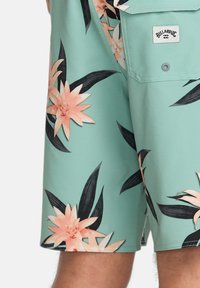 Mätovo zelené plavky s kvetinovým vzorom, pričom obsahujú ružové kvety a čierne listy. Majú náprsné vrecko s označením značky.