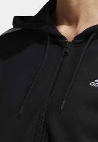 Närbild av en svart zip-up hoodie med huva, dragsnören och en vit Adidas-logotyp på bröstet.