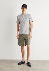 Γκρι κοντομάνικο πόλο πουκάμισο, ελκυστικά πράσινα σορτς με σχέδια και μαύρα παντόφλα. Μοντέλο φορώντας ένα μαύρο καπέλο με άσπρο κείμενο.