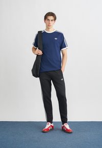 Námornícky modré tričko s bielymi prúžkami, skombinované s čiernymi zužujúcimi sa teplákmi so bočnými prúžkami a vyšitým logom; červené športové topánky.