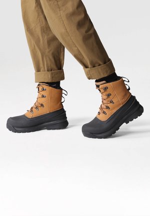 Braune wasserdichte Stiefel mit schwarzen Gummisohlen. Zu den Merkmalen gehören Metallösen, Schnürsenkel und ein gepolsterter Schaft. Getragen mit hochgekrempelten khakifarbenen Hosen.