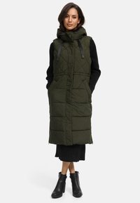 Dugi zeleni puffer prsluk s kapuljačom, s kvadratnim dizajnom i podesivim trakama, nosi se preko crne haljine od rebrastog trikota. Crne čizme do gležnja.