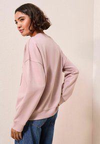 Felpa oversize rosa chiaro con colletto a girocollo e maniche lunghe, abbinata a jeans di denim, su uno sfondo neutro.