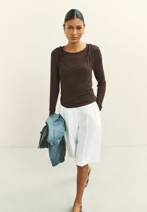 Nő szűk barna hosszú ujjú felsőben és fehér, térdig érő rövidnadrágban, egyik kezében kék dzsekit tartva, előre sétál egy egyszínű háttér előtt.