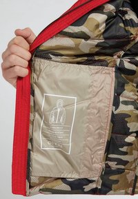 Padded Jacke mit rotem Außenmaterial und camo-farbenem Innenfutter. Enthält eine sandfarbene Innentasche mit Pflegehinweisen und wasserabweisenden Materialien.