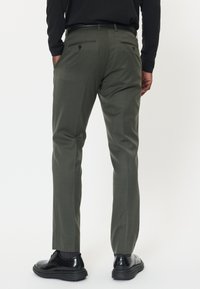 Olivgröna byxor med skräddarsydd passform, med bakfickor och en smidig textur. I kombination med svarta skor, står på en enkel bakgrund.