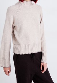 Pull en laine beige avec col roulé côtelé et poignets. Design court avec coutures surélevées et texture légère. Porté avec un pantalon foncé.