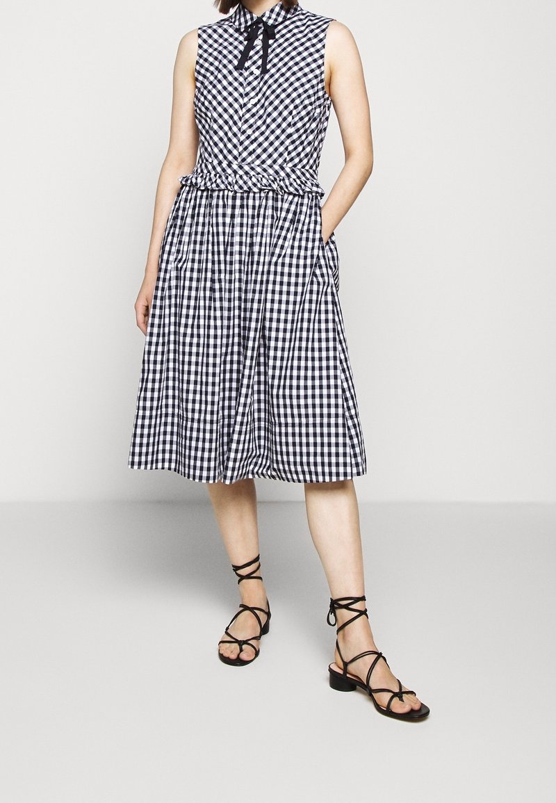 Femme portant une robe sans manches à carreaux noirs et blancs avec un col nœud papillon, des poches et des sandales noires à brides et talons bas, debout sur un sol gris.