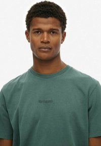 Zielona bawełniana koszulka z okrągłym dekoltem i krótkimi rękawami, z subtelnym haftowanym logo "SUPERDRY" na piersi w dopasowanym kolorze.