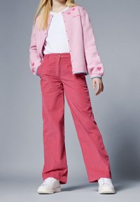 Cardigan rosa chiaro con motivi a cuore, chiusura a bottoni, abbinato a pantaloni rosa a vita alta e gamba larga, e sneaker bianchi.