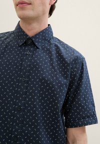 Short-sleeve shirt in navy blue cotton with a small white and green geometric pattern, featuring a collared neck and button closure.