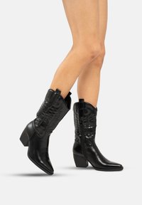 Botas de cowboy de cuero negro con punteras puntiagudas y tacón cuadrado, que presentan costuras decorativas y lengüetas laterales para ponerlas y quitarlas fácilmente.