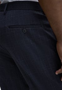 Marinefarbene Anzughose mit einem dezenten strukturierten Muster. Besitzt eine Gesäßtasche mit einem schwarzen Knopf und eine glatte Oberfläche. Maßgeschneiderte Passform.