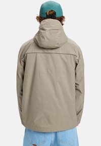 Lichtbeige jas met een capuchon, met een gladde structuur en een reflecterende accent over de schouder. Gekleed met lichtblauwe jeans.