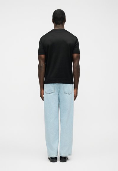 Άνδρας που φοράει μαύρο πουκάμισο με κοντά μανίκια, ανοιχτό μπλε τζιν με φαρδιά πόδια και μαύρα παπούτσια, στέκεται με την πλάτη γυρισμένη σε λευκό φόντο.