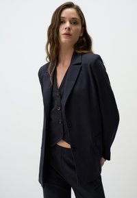 Blazer a righe navy con revers a notched, due tasche frontali e chiusura con un solo bottone, abbinato a un gilet coordinato.
