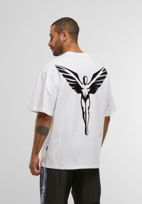 Mann med kort hår og tatovering på armen, iført hvit oversized T-skjorte med stor svart abstrakt vinget figur på ryggen og svarte bukser.