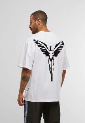 Mann med kort hår og tatovering på armen, iført hvit oversized T-skjorte med stor svart abstrakt vinget figur på ryggen og svarte bukser.