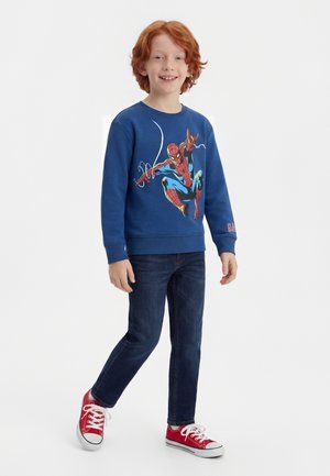 Red-haired boy smiling, wearing a blue Spider-Man sweatshirt, dark jeans, and red sneakers, standing with one foot forward on white background.