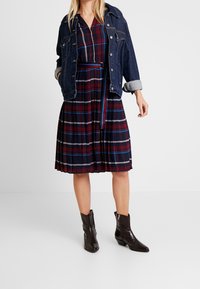 Kobieta w niebieskiej i czerwonej sukience w kratkę z cienkim paskiem, ciemnogranatowej kurtce dżinsowej na ramionach i czarnych botkach za kostkę na niskim obcasie.