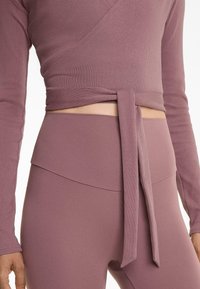 Rebrasti, dugih rukava, wrap top u ljubičastoj boji s vezanjem u struku. Visok struk, odgovarajuće tajice s višeslojnim pojasom. Glatka tekstura.
