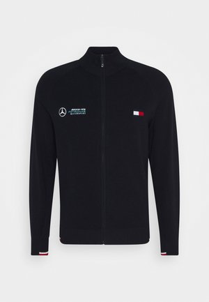 Czarna kurtka na pełny zamek z logo Mercedes AMG Petronas Motorsport po lewej stronie klatki piersiowej oraz małą, czerwoną, białą i niebieską flagą po prawej stronie klatki piersiowej.