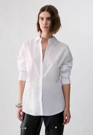 Jeune femme aux cheveux bruns courts porte une chemise blanche ample à boutons et un pantalon en cuir noir avec poches sur un fond clair uni.