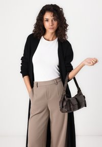 Brun skinnveske med flere lommer, holdt av en person i svart cardigan og beige høytlivs bukser, satt mot en hvit bakgrunn.